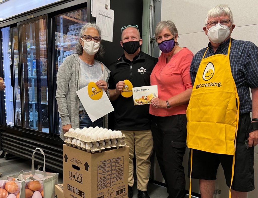 four people stand around cases of eggs and egg totes, inside a food bank building.